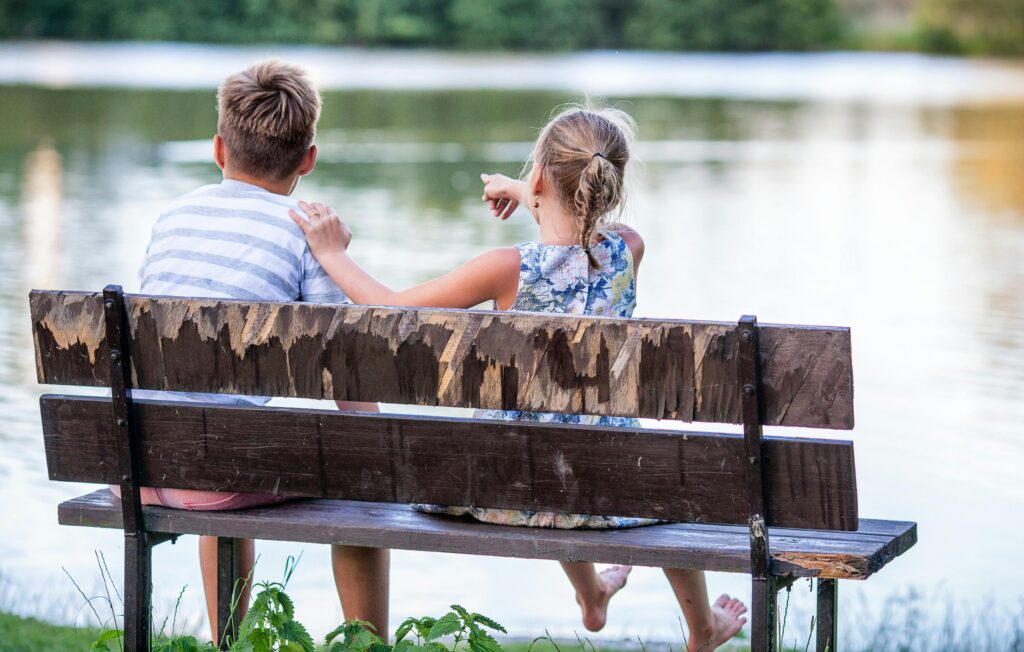 Two children sitting on a bench by a lake enjoying the view on a sunny day.
